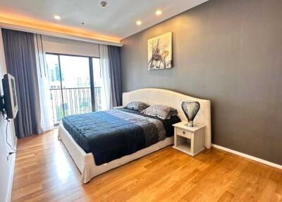 Spacious bedroom with wooden flooring, modern furniture, large window with curtains, and wall-mounted TV.