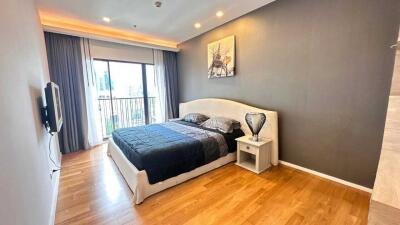 Spacious bedroom with wooden flooring, modern furniture, large window with curtains, and wall-mounted TV.