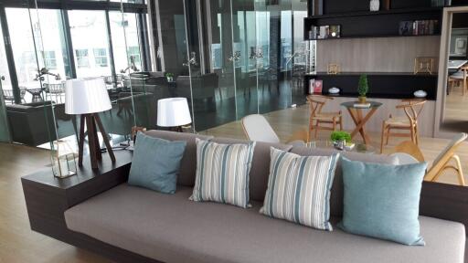 Modern living room with a grey sofa, decorative pillows, glass walls, and a dining area in the background