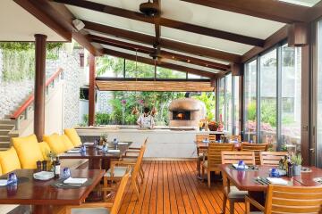 Spacious outdoor dining area with wooden tables, chairs, and large windows.