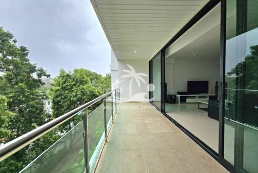 Spacious balcony with glass railing and view of surrounding greenery, adjacent to a modern living room.