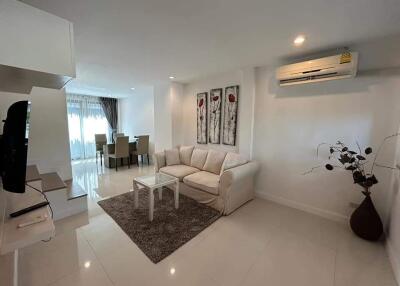 A modern living room with a white sofa, coffee table, wall-mounted TV, dining area, and air conditioning