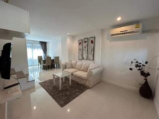 A modern living room with a white sofa, coffee table, wall-mounted TV, dining area, and air conditioning