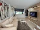 Modern living room with a beige sofa, glass coffee table, wall-mounted TV, and large window with gray curtains