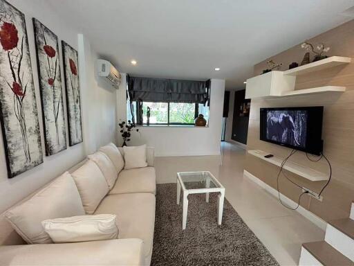 Modern living room with a beige sofa, glass coffee table, wall-mounted TV, and large window with gray curtains