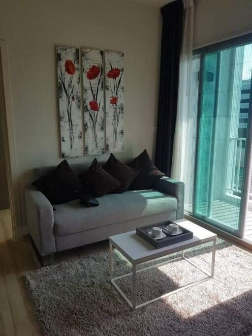 Modern living room with a grey sofa, decorative pillows, wall art with red flowers, coffee table with a tray, and large window with balcony view