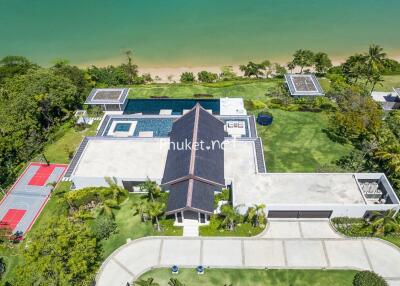 Aerial view of a luxurious beachfront property with a swimming pool, tennis court, and extensive green lawn.