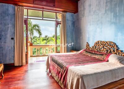 Spacious bedroom with large window overlooking greenery, featuring a wooden bed with intricate headboard and wooden furniture