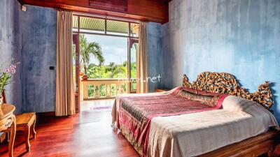 Spacious bedroom with large window overlooking greenery, featuring a wooden bed with intricate headboard and wooden furniture