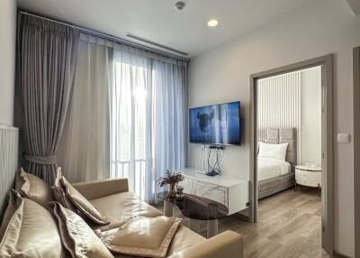 A modern living room with a beige sofa, a wall-mounted TV, and a view into the bedroom