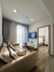 A modern living room with a beige sofa, a wall-mounted TV, and a view into the bedroom