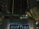 View of a modern high-rise building at night with illuminated 'RHYTHM' sign