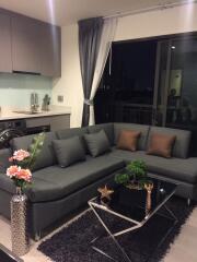 Modern living room with gray L-shaped sofa, decorative plants, glass coffee table, and kitchen area in the background