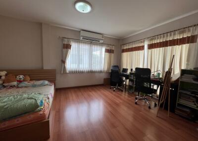 A spacious bedroom with wooden flooring, featuring a bed, a study area with two desks and chairs, bookshelves, and large windows with curtains.