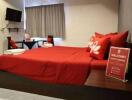 Bedroom with a double bed featuring red pillows and bedsheet, a wall-mounted TV, and a small seating area with a table and two chairs.