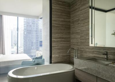 Modern bathroom with bathtub, sink, and large mirror, adjacent to a bedroom with city view