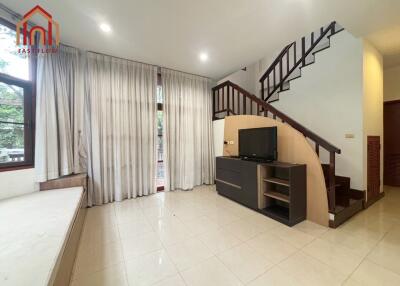 Spacious living room with tiled flooring, a wooden staircase, large windows with curtains, and a TV cabinet.