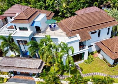 Large modern house with brown tile roof, multiple levels, lush greenery, and palm trees.