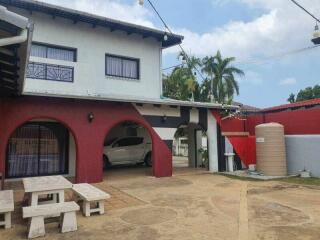 Exterior view of a building with an attached carport, outdoor seating, water tank, and palm trees.