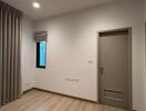 Empty bedroom with light wooden flooring, a small window with curtains, and a closed door