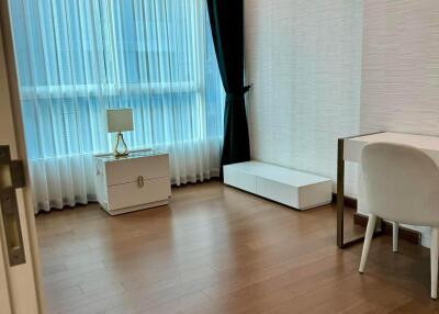 Modern bedroom with wooden floors, a large window with white and black curtains, a white desk and chair, a white stand, and a small white cabinet with a lamp