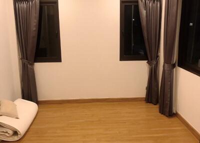 Empty bedroom with wooden flooring, two windows with curtains, air conditioning unit, and folded bedding on the floor.