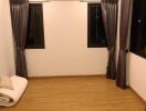 Empty bedroom with wooden flooring, two windows with curtains, air conditioning unit, and folded bedding on the floor.