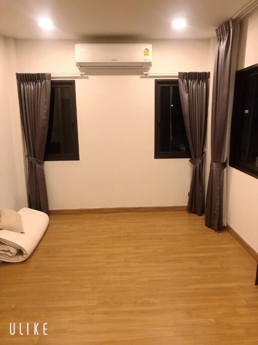 Empty bedroom with wooden flooring, two windows with curtains, air conditioning unit, and folded bedding on the floor.