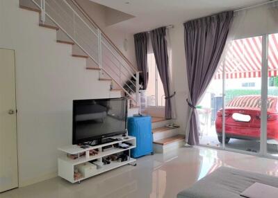 Living room with stairs to upper floor, a TV on white stand, large windows with curtains, and a view of a parking area