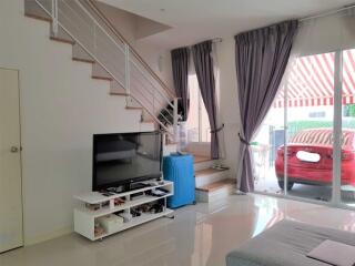 Living room with stairs to upper floor, a TV on white stand, large windows with curtains, and a view of a parking area