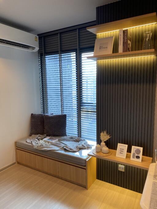 Cozy corner in a modern living area with storage bench, air conditioning, shelf with books and decor, and large window with blinds