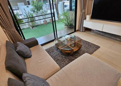 Modern living room with large sofa, glass coffee table, and entertainment center, adjacent to a patio with greenery.