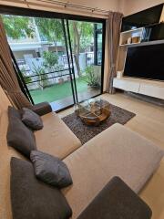 Modern living room with large sofa, glass coffee table, and entertainment center, adjacent to a patio with greenery.