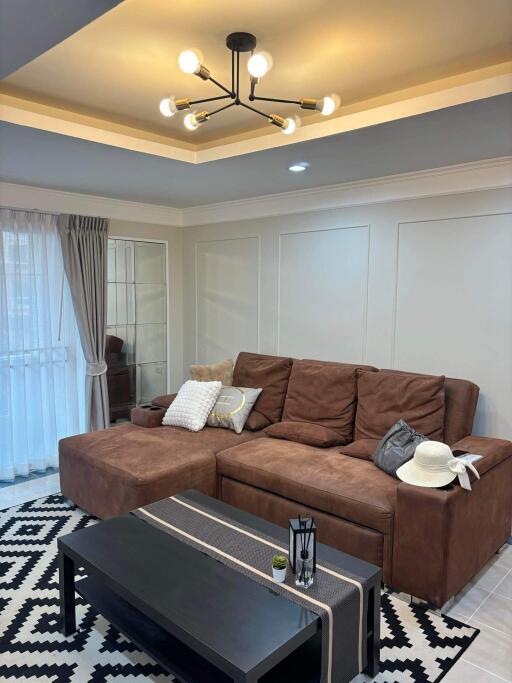 Modern living room with brown sectional sofa, black coffee table, stylish light fixture, and natural light from large windows.