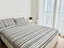 Bedroom with striped bedspread and bedside table next to a large window with sheer curtains