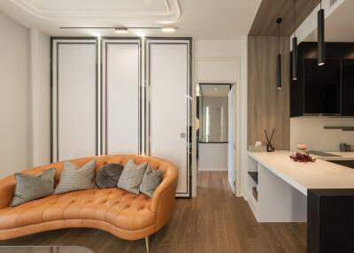 Modern living room with a curved, orange leather sofa, decorative cushions, adjacent to a kitchen area with a white countertop and black hanging lights