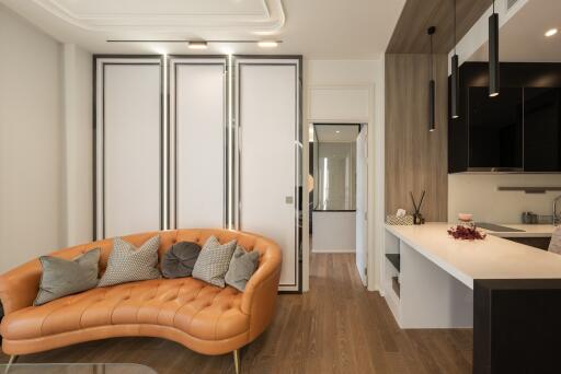 Modern living room with a curved, orange leather sofa, decorative cushions, adjacent to a kitchen area with a white countertop and black hanging lights