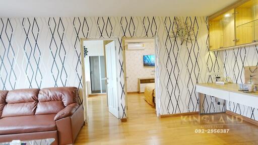 A cozy living room with a leather sofa, wooden flooring, patterned wallpaper, built-in shelves and desk, and a view into an adjacent bedroom