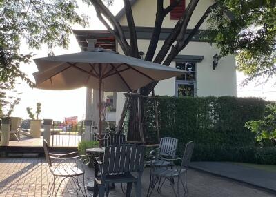 Outdoor seating area with table and chairs under an umbrella, next to a house surrounded by trees.