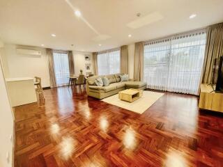 Spacious living room with wooden flooring, large windows, sofa, coffee table, dining area, air conditioning unit, and ample natural light.