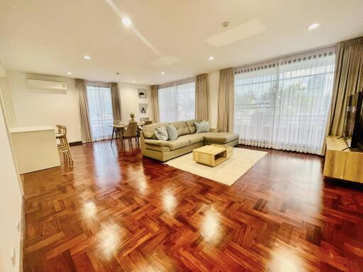 Spacious living room with wooden flooring, large windows, sofa, coffee table, dining area, air conditioning unit, and ample natural light.