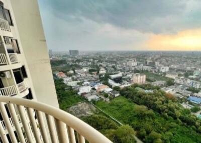 View from a high-rise apartment balcony showing a cityscape with greenery and buildings under a cloudy sky