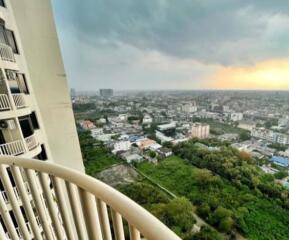 View from a high-rise apartment balcony showing a cityscape with greenery and buildings under a cloudy sky