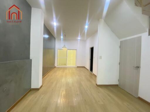 Spacious living room with wooden flooring, ceiling lights, and adjacent hallway.