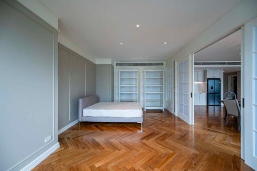 Spacious bedroom with wooden flooring and large bed, featuring built-in shelves and a view towards the kitchen area.