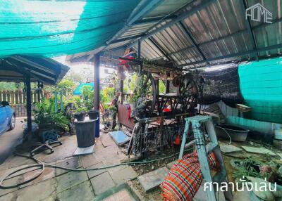 Messy outdoor workspace under a corrugated metal roof with various tools, construction materials, and plants.