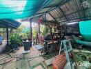 Messy outdoor workspace under a corrugated metal roof with various tools, construction materials, and plants.