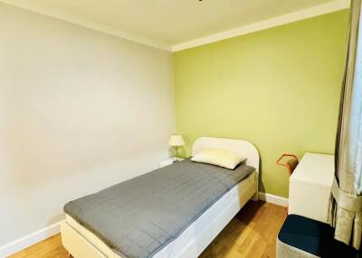 Single bed in a small, well-lit bedroom with green and white walls, wooden floor, and minimalist furniture