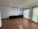 Spacious living room with wooden flooring, large windows with curtains, and air conditioning.