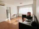 Spacious living room with black leather sofa, window with curtains, and wooden floor